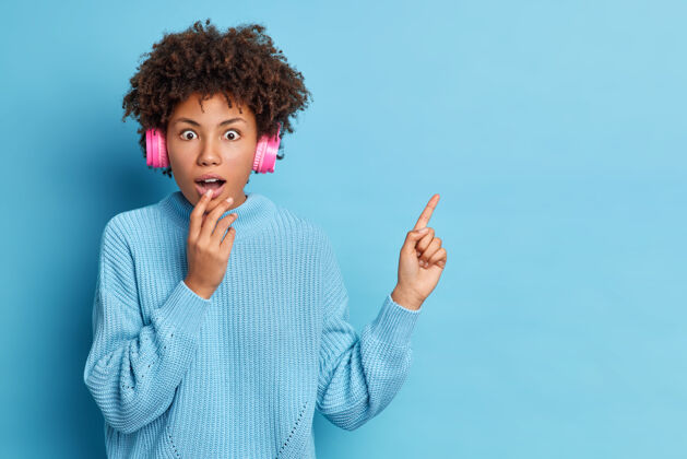 穿震惊的年轻非洲裔美国妇女惊奇地张大嘴巴戴着街头耳机听最喜欢的音乐在复印空间表明穿着针织毛衣卷发手指手势