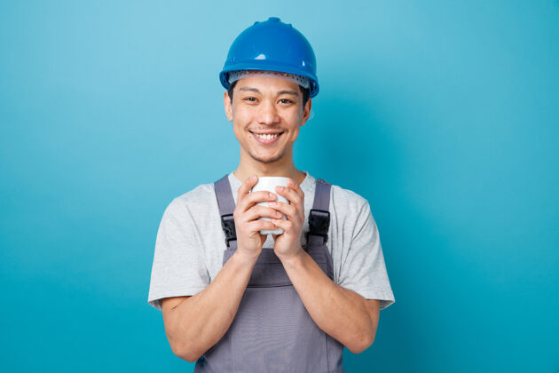 建筑微笑的年轻建筑工人戴着安全帽 穿着制服 双手捧着茶杯戴着蓝色头盔