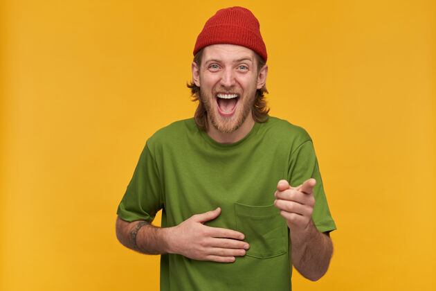 休闲正面的 成年男性的肖像 金色的发型和胡须穿着绿色的t恤和红色的帽子有纹身你笑得很厉害隔着黄墙隔离笑装束积极