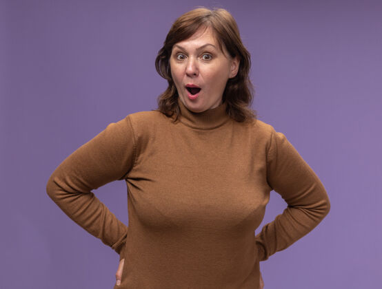 中等穿着棕色高领毛衣的中年妇女站在紫色的墙上年龄女人站着