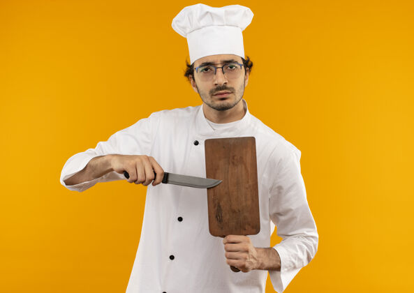 板自信的年轻男厨师穿着厨师制服 戴着眼镜 手里拿着砧板和刀切割制服刀