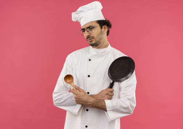 困惑看着身边困惑的年轻男厨师穿着厨师制服 戴着眼镜拿着勺子和煎锅烹饪穿眼镜