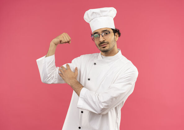 烹饪年轻的男厨师穿着厨师制服 戴着眼镜做着坚强的手势制服男性男士