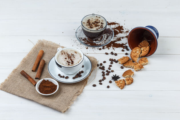 早晨一套咖啡豆 磨碎的咖啡 曲奇饼 肉桂棒和咖啡杯在木制和布袋背景高角度视图肉桂卡布奇诺