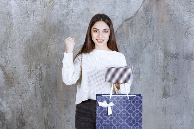 人体模特一个穿白衬衫的女孩手里拿着一个银色的礼盒和一个蓝色的购物袋 并显示出积极的手势员工快乐女销售员
