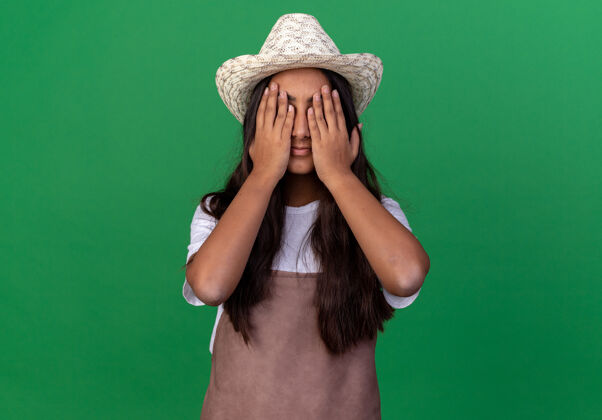 花园年轻的园丁女孩围着围裙戴着夏令帽闭着眼睛站在绿色的墙上关闭帽子年轻的