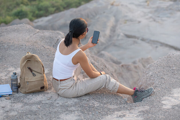 人年轻女子拿着手机坐着夕阳下的高山旅游小道背包徒步旅行步行