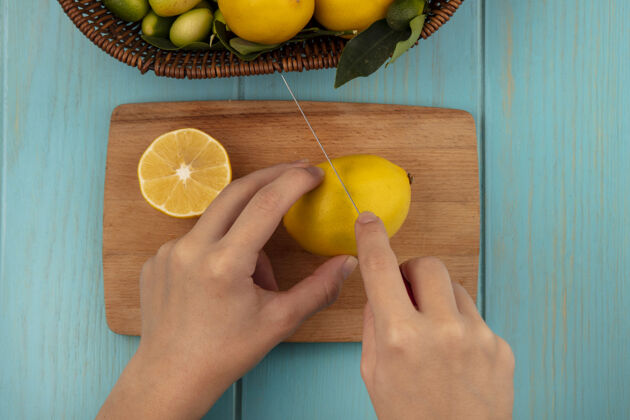 切女性用刀在木制厨房板上切割新鲜柠檬的手俯视图 蓝色木制表面上的桶上有金盏花和柠檬等水果饮食配料厨房