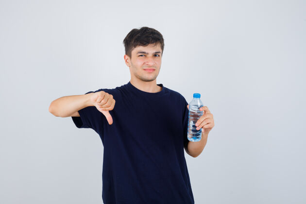 男人一幅年轻人拿着一瓶水的肖像 穿着黑色t恤 拇指朝下 看着不高兴的前视图自信愤怒成人
