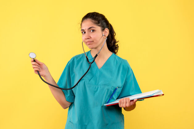 持有正面图穿着制服的年轻女医生拿着听诊器和剪贴板在黄色背景上诊所剪贴板医学