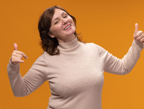 请穿着米色套头衫的中年妇女高兴地指着自己站在橘色的墙上 快乐而愉快中等年龄高领毛衣