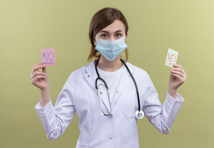 年轻年轻的女医生穿着医用长袍 戴着口罩和听诊器 拿着是和否的纸条 看着隔离的绿色墙壁抱着医生长袍