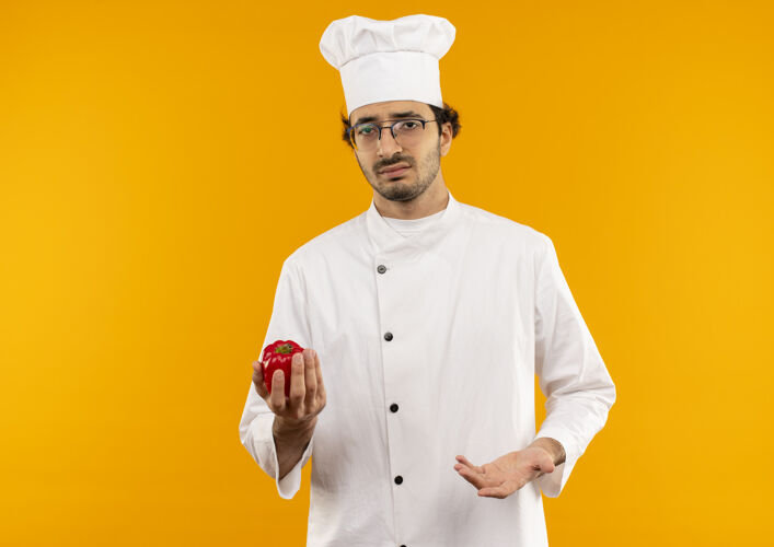 胡椒悲伤的年轻男厨师穿着厨师制服 戴着眼镜 手里拿着辣椒隔离在黄色的墙上悲伤眼镜持有