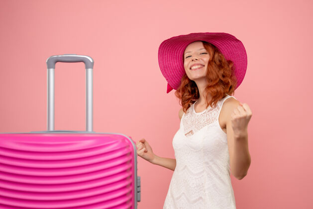 美丽年轻的女性游客与粉红色的帽子和粉红色的墙壁上的包的正面视图衣服快乐度假
