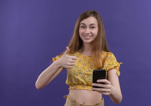 握着微笑的年轻女孩拿着手机 在孤立的紫色墙上竖起大拇指年轻人电话表演