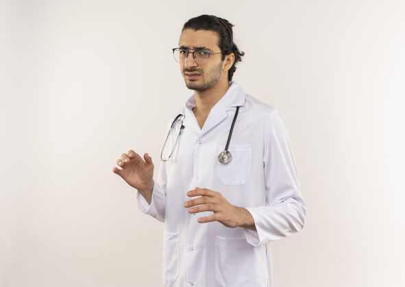 戴神经质的年轻男医生 戴着眼镜 穿着白色长袍 听诊器放在隔离的白墙上 留着复印空间男性听诊器年轻