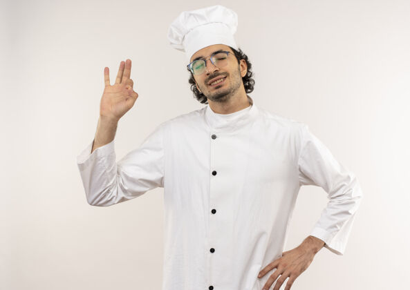 臀部年轻的男厨师闭着眼睛 穿着厨师制服 戴着眼镜 手放在白色墙壁上的屁股上 显得很高兴眼镜年轻手势