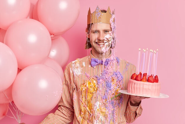 祝贺英俊开朗的男人站在脏兮兮的奶油摊上 拿着美味的草莓蛋糕庆祝生日 站在粉色的墙上 与节日属性隔离欢呼草莓抱持