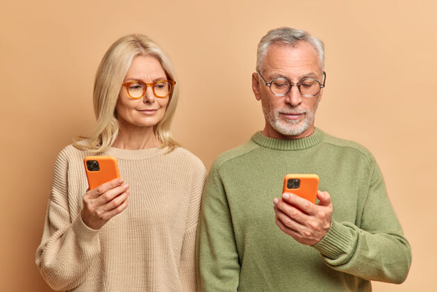 团结几位资深女士和男士使用现代智能手机 专注于展示阅读在线新闻 穿着休闲套头衫 隔着棕色的工作室墙壁老年人在一起眼镜