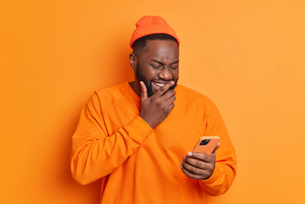 观看积极的男人在智能手机屏幕上咯咯地笑 在互联网上观看有趣的视频 或者在收到的信息上笑 穿着休闲明亮的衣服 隔离在橙色的墙上男性快乐人