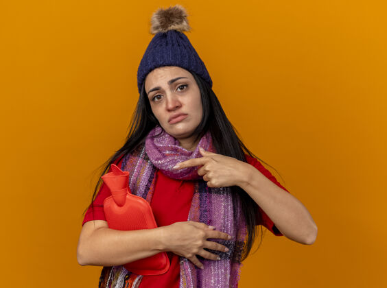 悲伤悲伤的年轻白种人生病的女孩戴着冬天的帽子和围巾拿着和指着孤立在橙色墙上的热水袋帽子炎热指尖