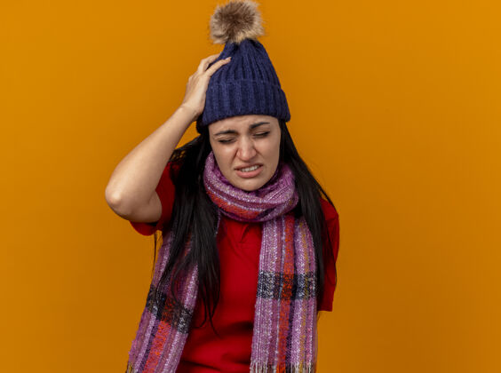 保持疼痛的年轻白种人生病的女孩戴着冬天的帽子和围巾 手放在头上 闭着眼睛 隔离在橙色的墙上 留着复制空间围巾头年轻