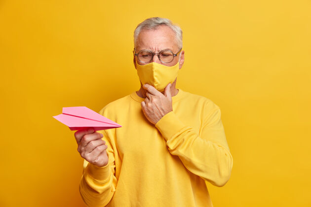 眼镜体贴的男性退休人员戴着眼镜水平镜头注视着纸飞机有着严重的膨胀思考着如何克服疾病在检疫时戴上防护口罩摆在室内纸单色扔
