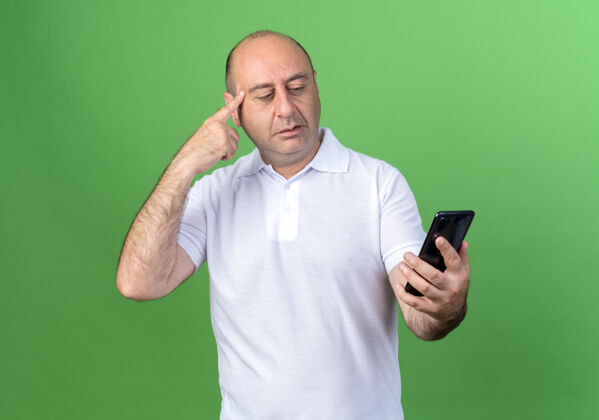 手指思想随意成熟的男人拿着手机看着手指放在太阳穴隔离在绿色的墙上男人太阳穴电话