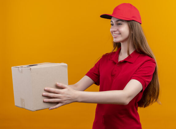 左侧微笑着的年轻送货女孩穿着红色制服 在孤立的橙色墙上 从左边的盒子里伸出来年轻侧线制服
