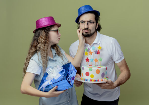 男人悲伤的年轻夫妇戴着粉色和蓝色的帽子 女孩拿着礼盒 男孩抱着脸颊 男孩拿着生日蛋糕 隔离在橄榄绿的墙上悲伤年轻蛋糕