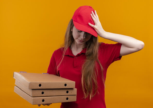 年轻紧张的年轻送货女孩身穿红色制服 手放在头上拿着包裹 闭着眼睛站在孤立的橙色墙上交货女孩头