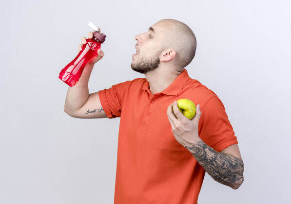 个人资料站在侧视图年轻的运动男子举行水瓶和苹果隔离在白色的墙壁上年轻苹果观点