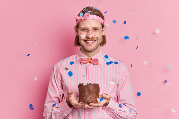成人快乐的成年男子庆祝在公司工作一年拿着小蛋糕庆祝同事们的祝贺微笑着高兴地戴着头带条纹衬衫和蝴蝶结科菲蒂落在他身上快乐生日五彩纸屑