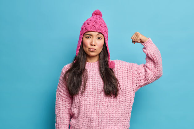 健康严肃的黑发女人举起手臂 露出肌肉自信而充满力量戴着针织套头衫粉色帽子感觉强壮健康隔离在蓝色的墙壁上手臂人漂亮