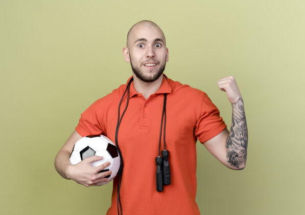 肩膀带着微笑的年轻运动型男子肩上扛着跳绳拿着球 在橄榄绿的墙上孤立地做着“是”的手势微笑握着运动