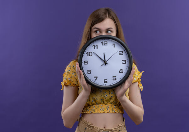 抱着年轻的女孩拿着钟 躲在它后面 看着左边的紫色墙壁上的复制空间时钟左边年轻