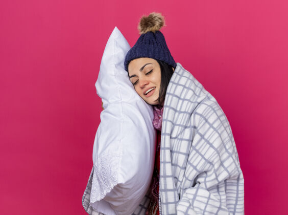 枕头微笑着的年轻白种人生病的女孩戴着冬天的帽子和围巾 裹着格子呢抱枕 把头放在上面 闭着眼睛 隔离在深红色的墙上 留着复制空间格子拥抱围巾