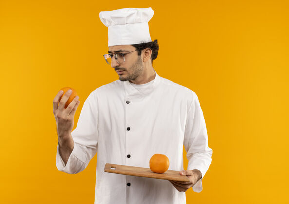 烹饪想着年轻的男厨师穿着厨师制服 戴着眼镜 拿着橘子看着隔离在黄色墙上的砧板思考男性持有