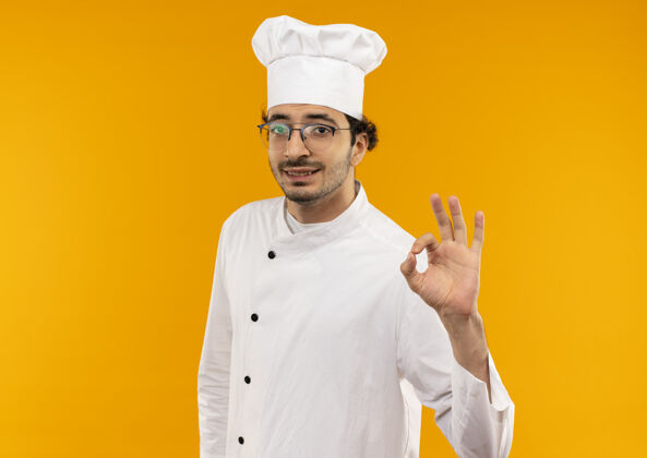 好的年轻的男厨师穿着厨师制服 戴着一副眼镜 在黄色的墙上显示出很好的姿势烹饪穿请
