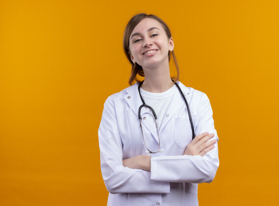 姿势面带微笑的年轻女医生穿着医用长袍和听诊器 以封闭的姿势站在隔离的橙色墙壁上 有复印空间年轻听诊器闭嘴