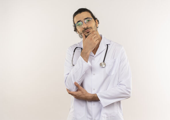 年轻年轻的男医生戴着眼镜 穿着白色长袍 听诊器 手放在下巴上 隔着一堵白色的墙 留着复印空间思考长袍下巴