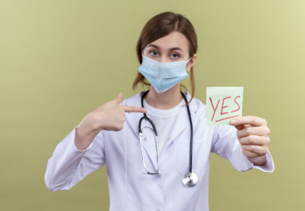 女人年轻的女医生穿着医用长袍 戴着口罩和听诊器 在隔离的绿墙上伸着“是”字条指着自己医学伸展指点