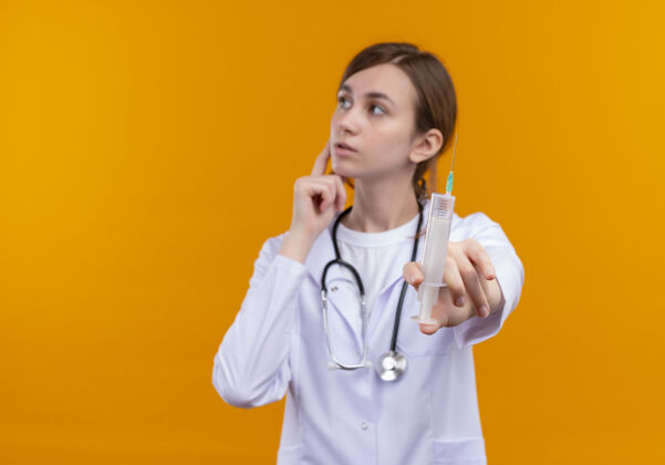 听诊器体贴的年轻女医生穿着医用长袍 手持听诊器和伸缩注射器 看着左侧隔离的橙色墙壁上的复印空间长袍思想医学