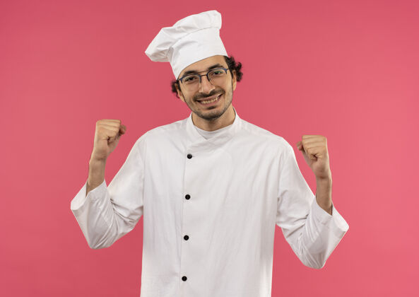 制服面带微笑的年轻男厨师穿着厨师制服 戴着眼镜 在粉红色的墙上显示“是”的手势手势微笑厨师