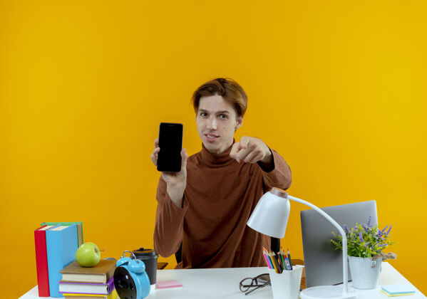 坐着高兴的年轻学生男孩坐在书桌旁 手里拿着学习工具 拿着电话 在黄色的墙上向你们展示孤立的手势拿着学校表演