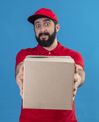 伸展印象深刻的年轻英俊的白人送货员穿着红色制服和帽子伸出纸箱对着镜头隔离在蓝色年轻印象穿着