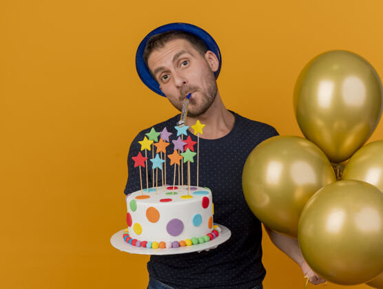 氦快乐英俊的白人男子戴着蓝色派对帽 手持氦气球和生日蛋糕吹口哨 橙色背景上有复制空间气球哨子蛋糕