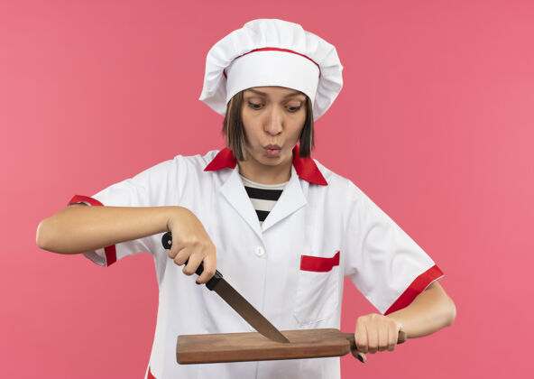 印象穿着厨师制服的年轻女厨师用刀抚摸着砧板 看着它孤立的粉红色板切割粉