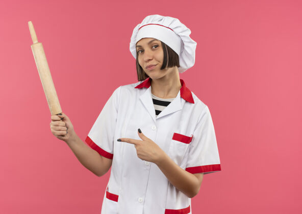 厨师自信的年轻女厨师 身着厨师制服 手拿擀面杖 指着粉红色的擀面杖持有烹饪粉