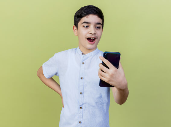 手机惊讶的年轻白人男孩拿着手机看着手放在腰上孤立在橄榄绿的背景与复制空间抱着保持手